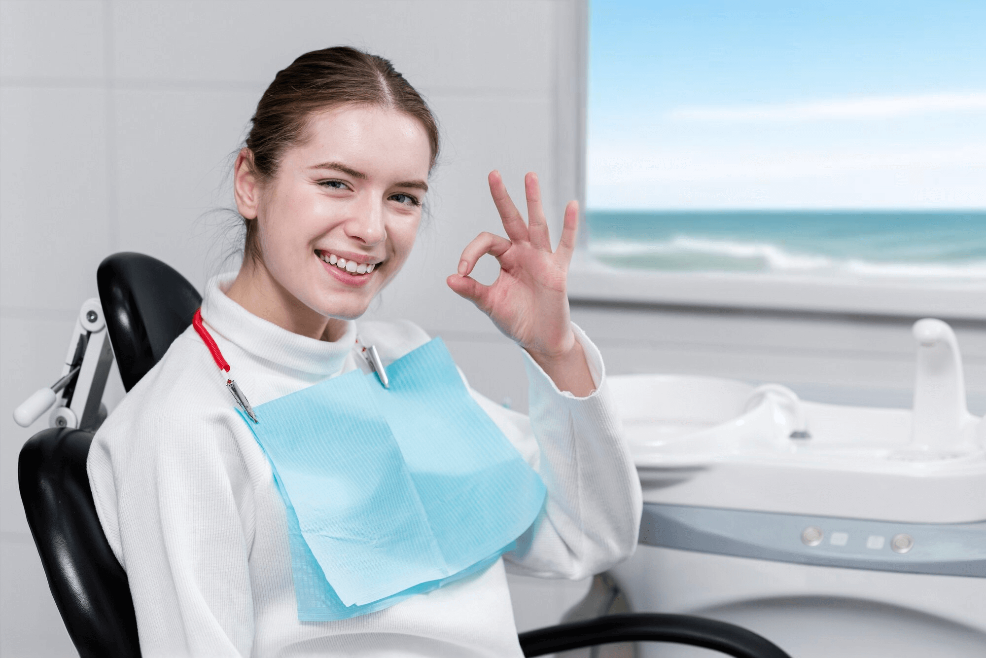 A smiling patient sitting in a modern dental clinic chair, with visible dental tools and a dentist in the background. Behind the patient, a large window reveals a scenic Mexican beach with palm trees, clear blue skies, and turquoise waters.